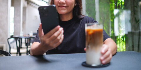 Closeup image of a woman holding a glass of iced latte coffee while using mobile phone in cafe