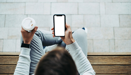 Person holding a phone with a blank screen and a coffee cup while sitting on a wooden bench