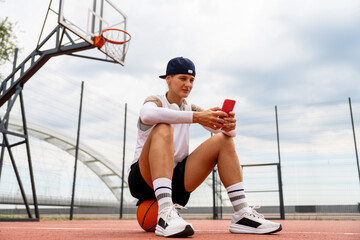 Female athlete sitting on basketball using smartphone on street court