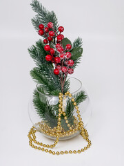 New Year's composition in a transparent round vase: a fir tree branch with red berries and gold beads on a white background