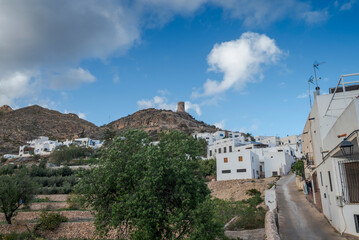The Atalaya de Nijar, a historic watchtower in Almeria, Spain, stands atop a rocky hill. The structure features a cylindrical design, surrounded by sparse greenery under a bright, partly cloudy sky