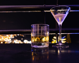 Two cocktail glasses with drinks on a rooftop bar railing at night overlooking the lights of a city in the background