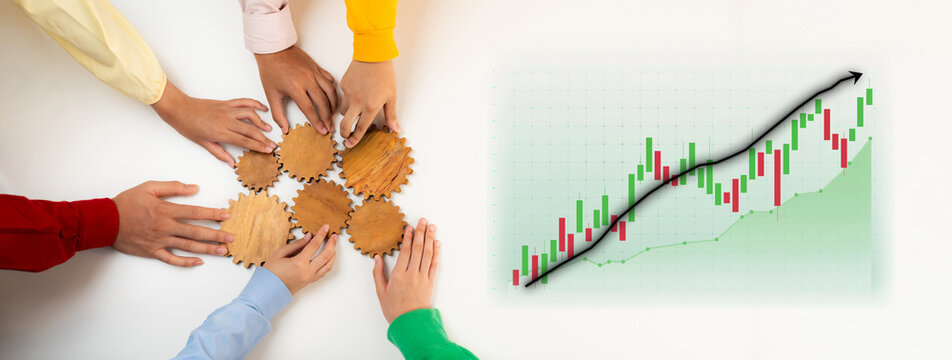 Diverse team gathers hands around wooden gears, symbolizing cooperation and strategy amidst financial growth chart, representing dynamic business environment. Axiom