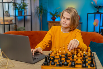 Caucasian teenager boy playing online chess on laptop app at home. Using a real chessboard with pieces on table, thoughtfully thinking and moving pieces while sitting comfortably on couch by table