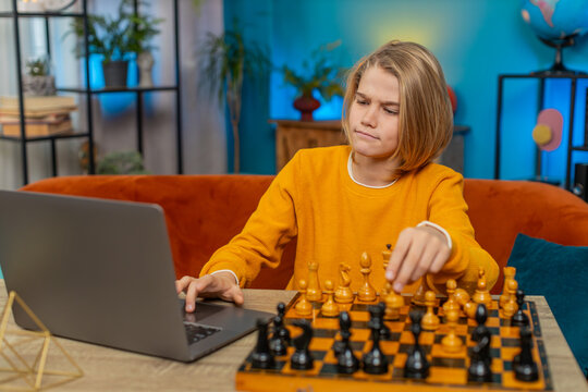 Caucasian teenager boy playing online chess on laptop app at home. Using a real chessboard with pieces on table, thoughtfully thinking and moving pieces while sitting comfortably on couch by table