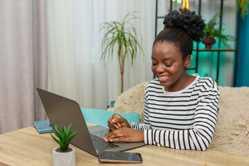 African American woman analyzing cryptocurrency data at home. Viewing charts on laptop, typing notes, checking rates on smartphone. Focused expression, working indoors, financial analysis setting.