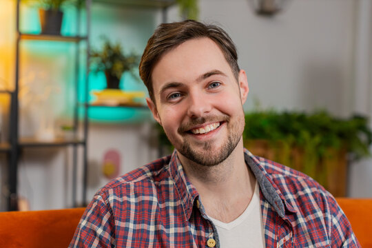 Portrait of happy Caucasian man sitting on sofa looking at camera and smiling at home. Young guy in casual shirt having attractive appearance enjoying leisure time relaxing in living room in apartment