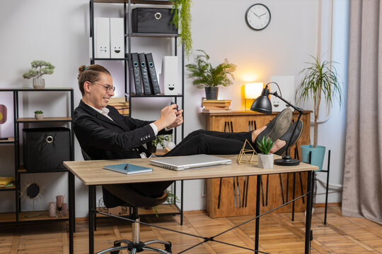Happy young man businessman enthusiastically playing shooter or racing video games on smartphone sitting at desk working from home office. Stylish man freelancer study, e-learning, remote job online.