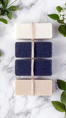 A top-down view of four rectangular bars of soap, each tied with natural twine, arranged vertically on a white marble surface, with green leaves and white bloss