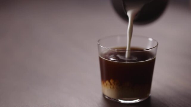 Slow motion milk pour into tumbler glass with coffee on walnut table