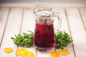 Bright red fruit drink in a glass pitcher with mint and dried orange slices on a wooden table