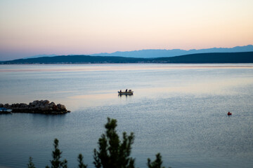 Small fishing boat with two silhouette figures sailing on the calm blue-purple Adriatic Sea at dawn, with hazy coastal mountains in the background, creating a mood of serenity and solitude