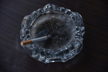 a lit cigarette in a clear glass ashtray with beautiful wisps of smoke on a textured wooden table...