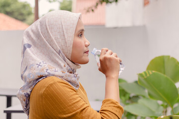 An Asian Muslim woman drinking from a plastic water bottle. Outdoors.