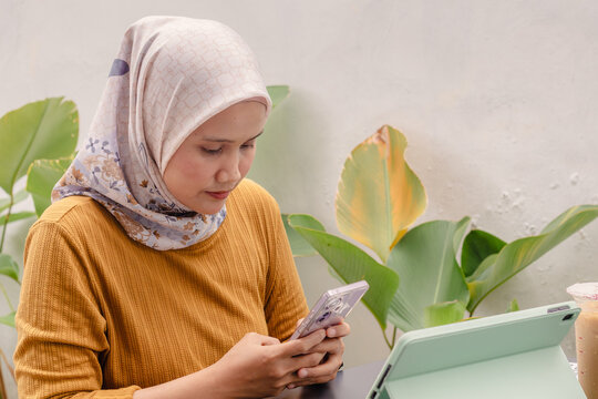 A young Indonesian Muslim woman working at an outdoor cafe using tablet and smartphone