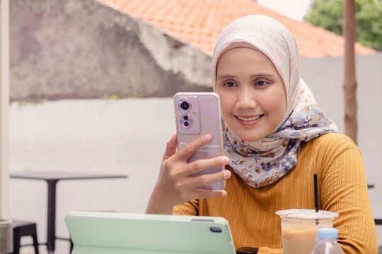 A young Asian Muslim woman receiving a video call from someone on her smartphone at an outdoor cafe