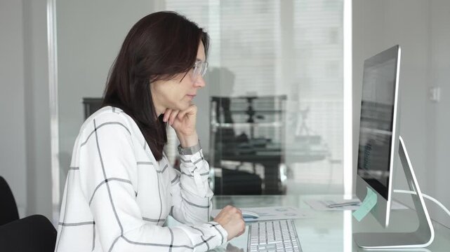Businesswoman in glasses working on computer, analyzing data and thinking in a modern office setup. Business people concept