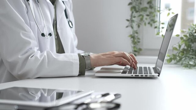Medical professional wearing white lab coat typing on laptop, documenting patient information with clinical precision at workstation. Medicine and health care concept