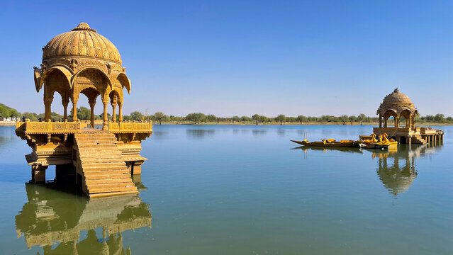 Ornate sandstone chhatri at Gadisar Lake in Jaisalmer, reflecting beautifully in the still water under a clear blue sky.