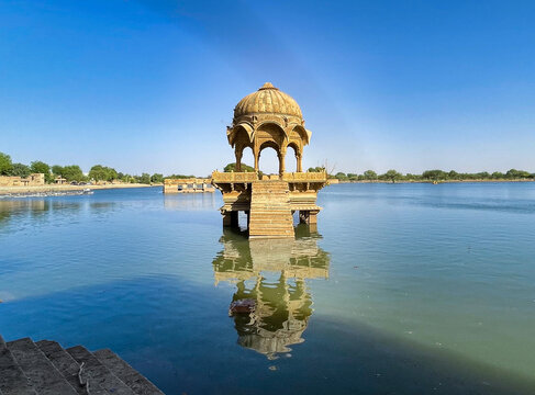 Ornate sandstone chhatri at Gadisar Lake in Jaisalmer, reflecting beautifully in the still water under a clear blue sky.