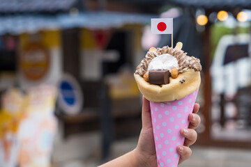 Hand holding a Japanese-style crepe filled with whipped cream, chocolate, and snacks with a small Japanese flag decoration