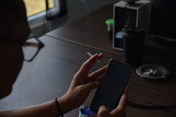 An Asian man wearing glasses is holding a cigarette and a cell phone in the office where he works in the morning.