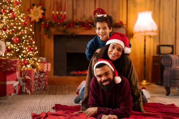Joyful African American family enjoying Christmas in warm living room, mother, father and boy wearing festive hats, lying on blanket in front of beautifully decorated tree