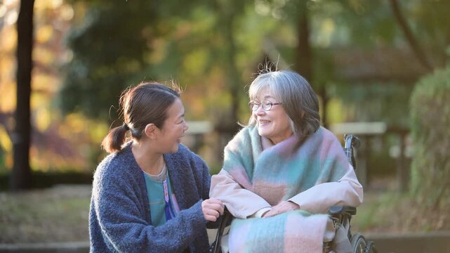 秋の紅葉で介護する女性と車椅子の高齢者の見つめ合う介護士のような人　落ち葉の前ボケあり