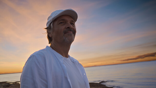 A middle-aged man in a white shirt and cap stands by the sea at sunset, embodying mindfulness and reflection. The serene coastal backdrop suggests a moment of mental wellbeing and escape.