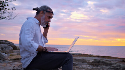 A focused digital nomad works on a laptop by the sea at sunset, embodying remote work freedom. The serene coastal setting highlights the blend of professional diligence and the allure of a portable.