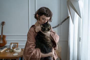 Sad melancholic woman holding cat close, seeking comfort, emotional support. Distressed girl cuddling beloved fluffy cat, standing near window at home. Empathy, connection between humans and animals.
