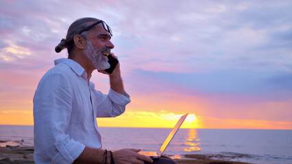 A bearded man with sunglasses on his head sits on a rocky shore at sunset, working on a laptop and...