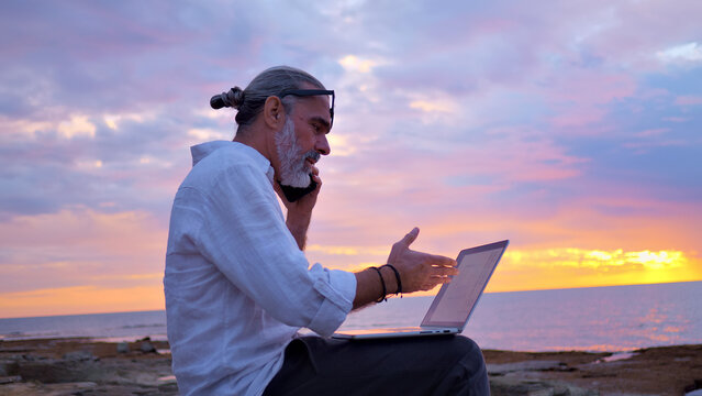 A bearded, middle-aged man in a white shirt works on a laptop on a rocky shore at sunset. The serene ocean and vibrant sky create a tranquil, remote setting, embodying digital nomadism.