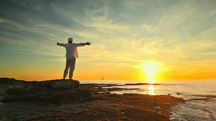 A person stands on a rocky shore at sunset, arms outstretched, embodying mindfulness and...