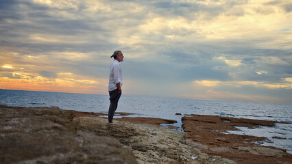 A man stands on rocky shores at sunset, gazing at the horizon. The serene ocean and pastel sky evoke mindfulness and reflection, capturing solitude and mental wellbeing.