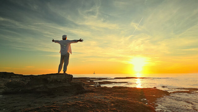 A person stands on a rocky shore at sunset, arms outstretched, embodying mindfulness and reflection. The serene ocean and golden sky evoke peace and escape, promoting mental wellbeing.