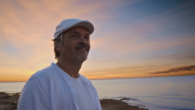 A middle-aged man in a white shirt and cap stands by the sea at sunset, embodying mindfulness and reflection. The serene coastal backdrop suggests a moment of mental wellbeing and escape.