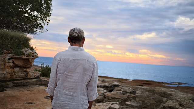 A person stands on rocky terrain, gazing at the ocean during sunset. The scene evokes mindfulness and reflection, capturing solitude and mental wellbeing.