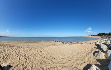 A sunlit beach stretches toward a calm sea and clear sky. Scattered rocks and the gentle curve of the coastline evoke an invitation to rest and contemplation.