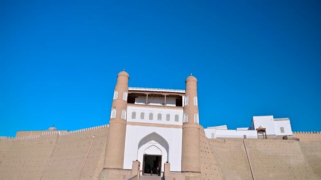 A smooth panning shot of the Ark of Bukhara in Uzbekistan, showing the curved outer walls, entrance area, and surrounding open courtyard under clear daylight.