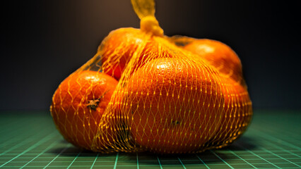 A net bag containing several bright oranges sits on a green cutting mat with a visible grid pattern. The dramatic lighting creates a vibrant contrast.