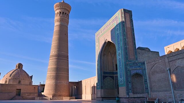 A wide view of the Kalan Minaret and mosque complex in Bukhara, Uzbekistan, showing the iconic brick tower and surrounding courtyard under bright daylight.