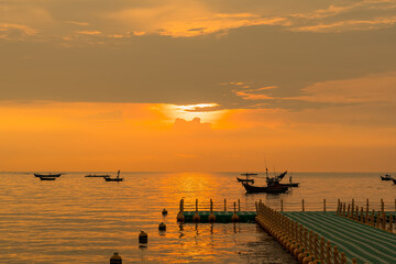 Floating dock extending into calm sea during golden sunrise with fishing boats on the horizon under a colorful sky