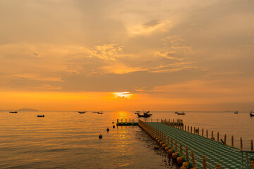 Floating dock extending into calm sea during golden sunrise with fishing boats on the horizon under a colorful sky