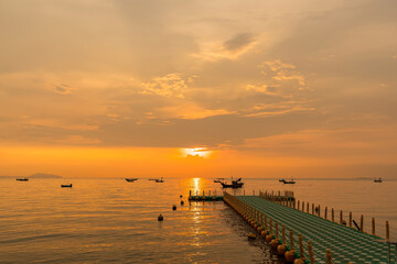 Floating dock extending into calm sea during golden sunrise with fishing boats on the horizon under a colorful sky