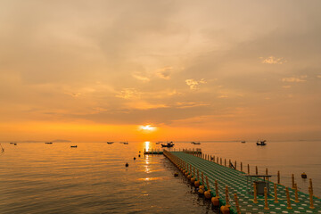 Floating dock extending into calm sea during golden sunrise with fishing boats on the horizon under a colorful sky