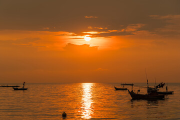 Fishing boats floating on calm sea during vibrant orange sunset with dock in foreground and golden reflections on water surface