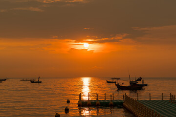 Fishing boats floating on calm sea during vibrant orange sunset with dock in foreground and golden reflections on water surface