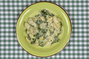 An overhead shot features gnocchi with creamy spinach sauce served on a green plate resting on a green and white checkered tablecloth