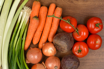 A top-down arrangement of fresh, raw vegetables including spring onions, carrots, onions, tomatoes, and beets, lying on a dark wooden surface